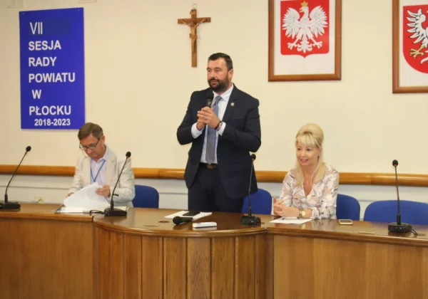 W sali konferencyjnej podczas ćwiczeń obronnych Zielony Jar 2019 mężczyzna w garniturze stoi i przemawia do mikrofonu przy stole, a po obu stronach siedzą dwie osoby; w tle banner z napisem "VII Sesja Rady Powiatu w Płocku 2018-2023" i herby.