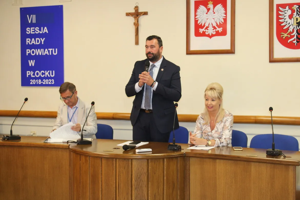 W sali konferencyjnej podczas ćwiczeń obronnych Zielony Jar 2019 mężczyzna w garniturze stoi i przemawia do mikrofonu przy stole, a po obu stronach siedzą dwie osoby; w tle banner z napisem "VII Sesja Rady Powiatu w Płocku 2018-2023" i herby.