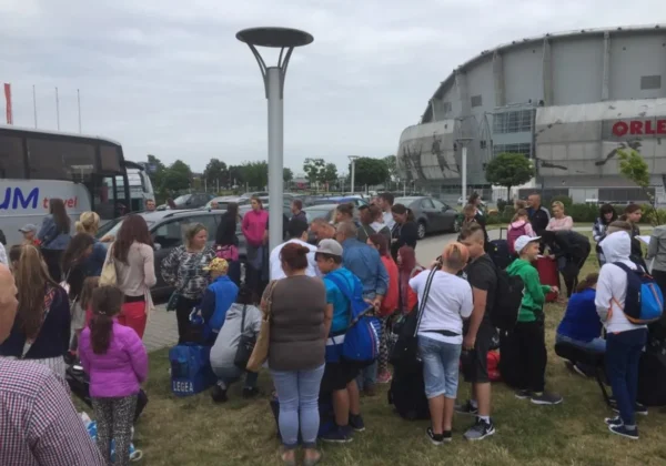 Grupa dzieci z opiekunami z plecakami i torbami stoi na parkingu obok autobusu przed dużym obiektem sportowym, przygotowując się do darmowych, 14‑dniowych kolonii nadmorskich dla mieszkańców powiatu płockiego.