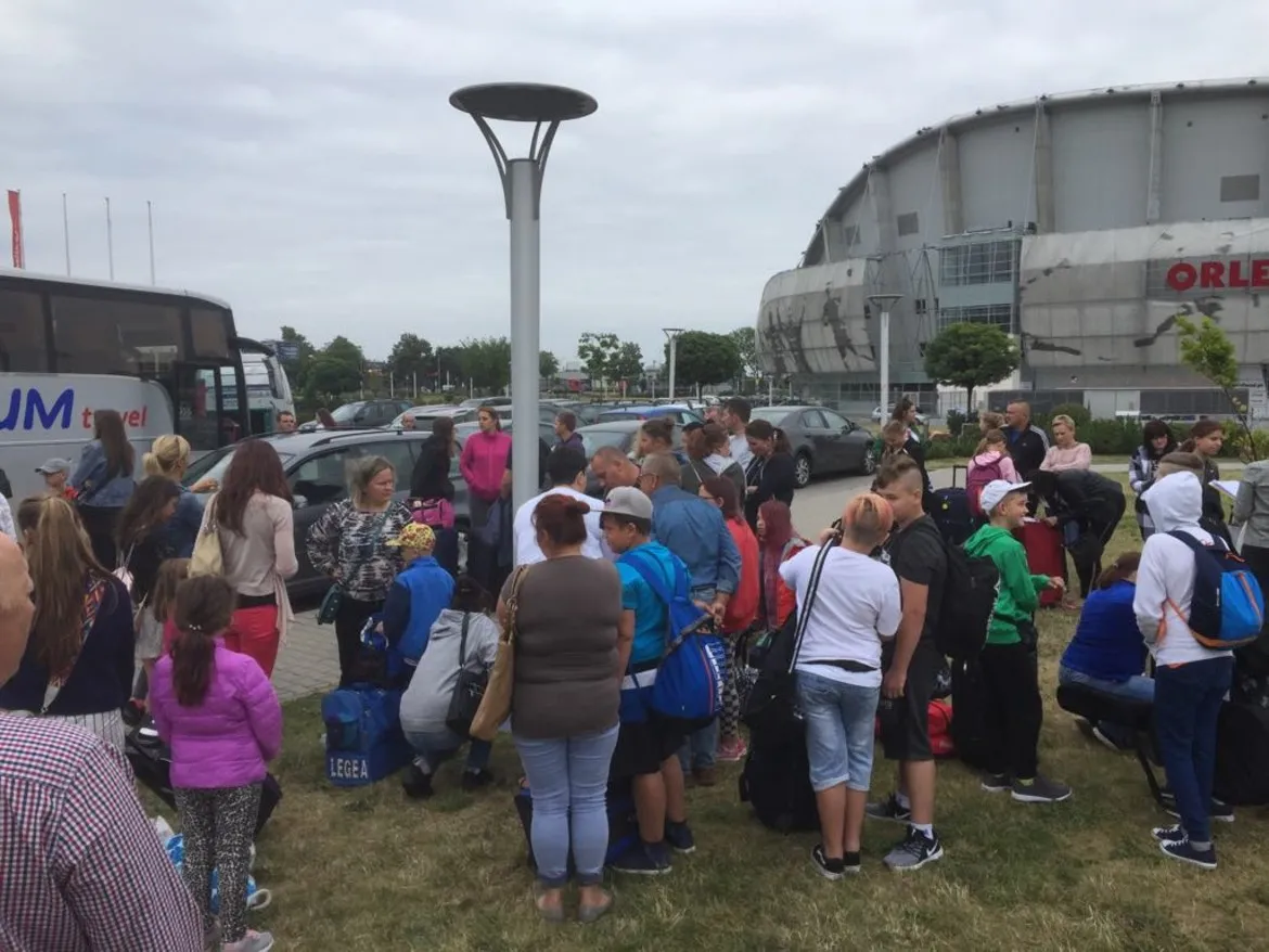 Grupa dzieci z opiekunami z plecakami i torbami stoi na parkingu obok autobusu przed dużym obiektem sportowym, przygotowując się do darmowych, 14‑dniowych kolonii nadmorskich dla mieszkańców powiatu płockiego.