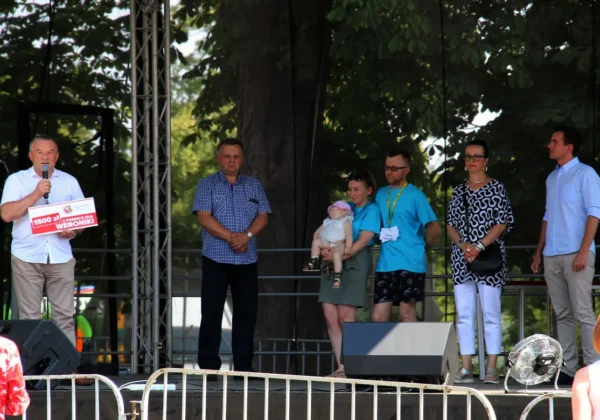 Osoby stoją na scenie podczas Pikniku Charytatywnego dla Weroniki; mężczyzna z mikrofonem trzyma plakat z informacją o wsparciu, a obok stoi rodzina z niemowlęciem.