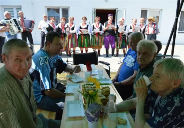 Mieszkańcy DPS w Koszelewie siedzą przy stole na świeżym powietrzu, obserwując występ zespołu Grzybowianki w tradycyjnych strojach ludowych na tle budynku.