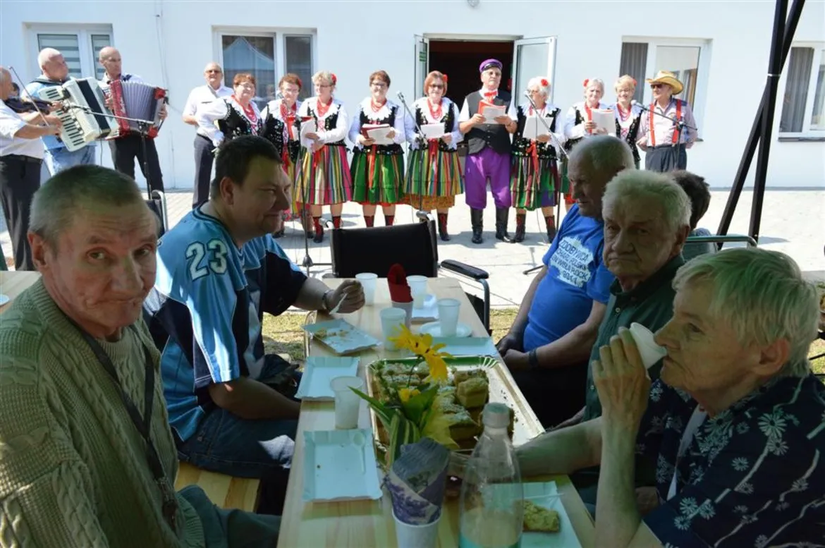 Mieszkańcy DPS w Koszelewie siedzą przy stole na świeżym powietrzu, obserwując występ zespołu Grzybowianki w tradycyjnych strojach ludowych na tle budynku.