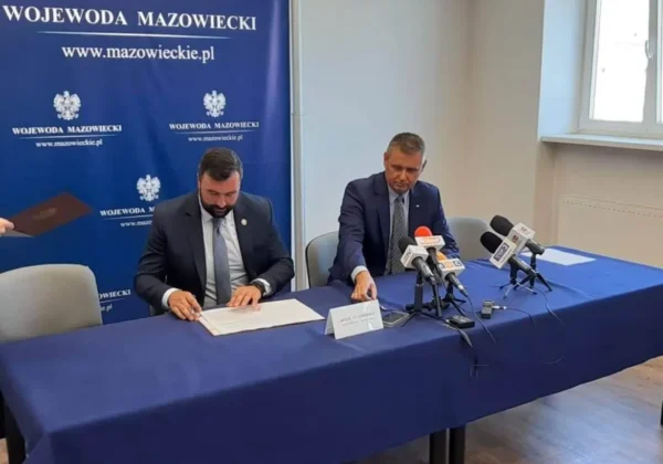 Dwóch mężczyzn w garniturach podpisuje dokumenty podczas konferencji prasowej dotyczącej nowej linii autobusowej łączącej Gminę Słubice z Miastem Płock, przy stole z mikrofonami i tłem z napisem Wojewoda Mazowiecki.