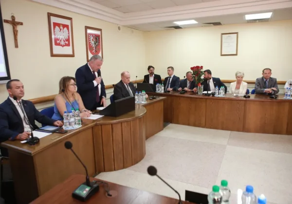 Uczestnicy IX Sesji Rady Powiatu siedzą przy półokrągłej drewnianej ławie w sali konferencyjnej, a jeden mężczyzna przemawia z mównicy; na stole widoczne są mikrofony, butelki z wodą i dokumenty, a w tle wiszą godła oraz krzyż.