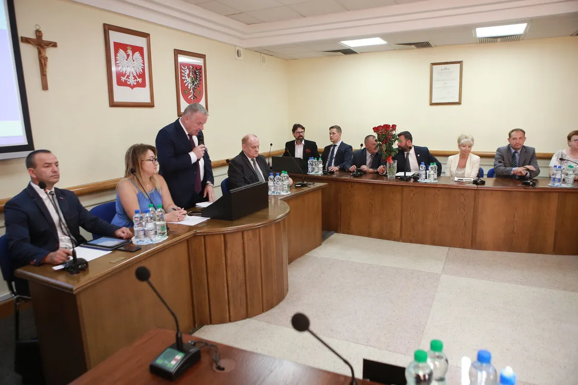 Uczestnicy IX Sesji Rady Powiatu siedzą przy półokrągłej drewnianej ławie w sali konferencyjnej, a jeden mężczyzna przemawia z mównicy; na stole widoczne są mikrofony, butelki z wodą i dokumenty, a w tle wiszą godła oraz krzyż.