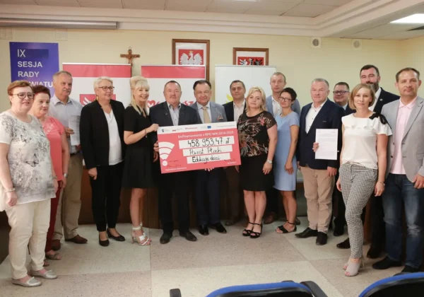 Grupa urzędników i samorządowców stoi w sali i prezentuje duży różowy czek z dofinansowaniem dla Powiatu Płockiego, w tle widoczne flagi i herby samorządowe.