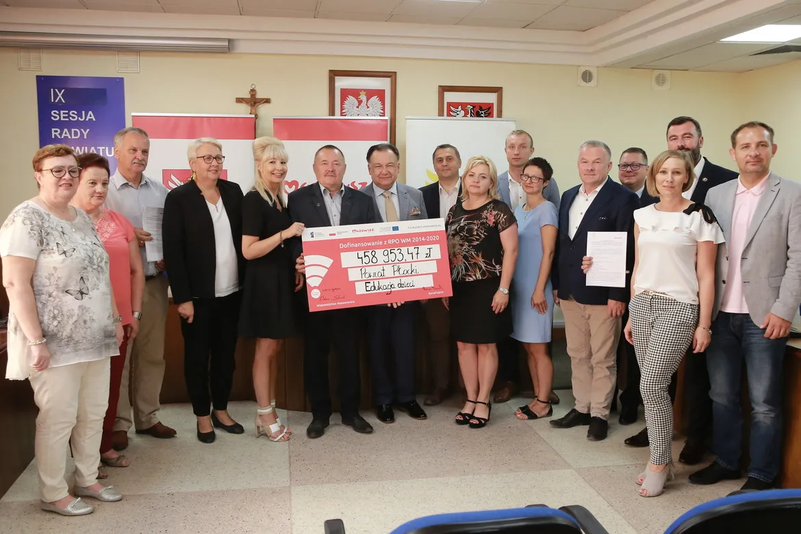 Grupa urzędników i samorządowców stoi w sali i prezentuje duży różowy czek z dofinansowaniem dla Powiatu Płockiego, w tle widoczne flagi i herby samorządowe.