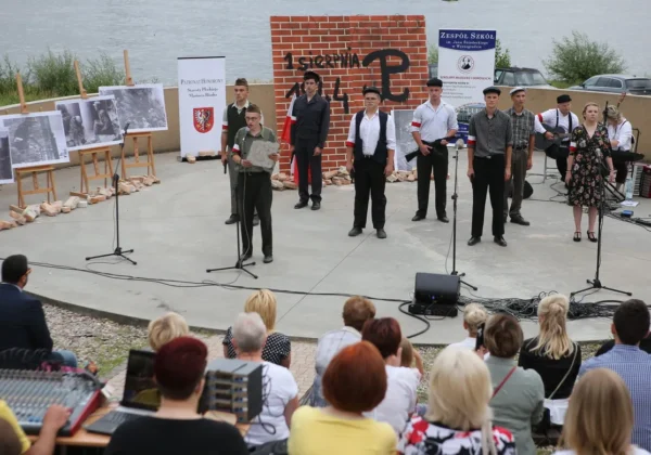 Scena plenerowa w Wyszogrodzie podczas powiatowych obchodów 75. rocznicy wybuchu Powstania Warszawskiego; grupa młodych wykonawców stoi przed mikrofonami, w tle tablica z napisem „1 sierpnia 1944” i czarno-białe fotografie na wystawie.