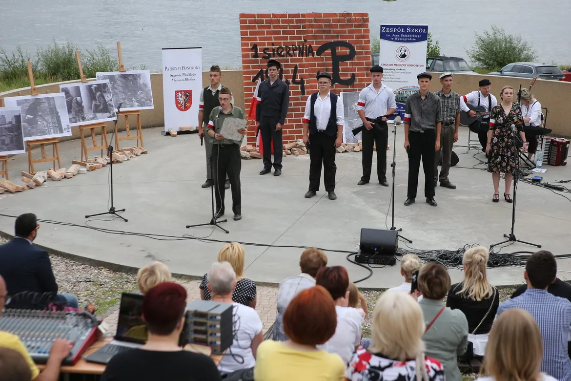 Scena plenerowa w Wyszogrodzie podczas powiatowych obchodów 75. rocznicy wybuchu Powstania Warszawskiego; grupa młodych wykonawców stoi przed mikrofonami, w tle tablica z napisem „1 sierpnia 1944” i czarno-białe fotografie na wystawie.