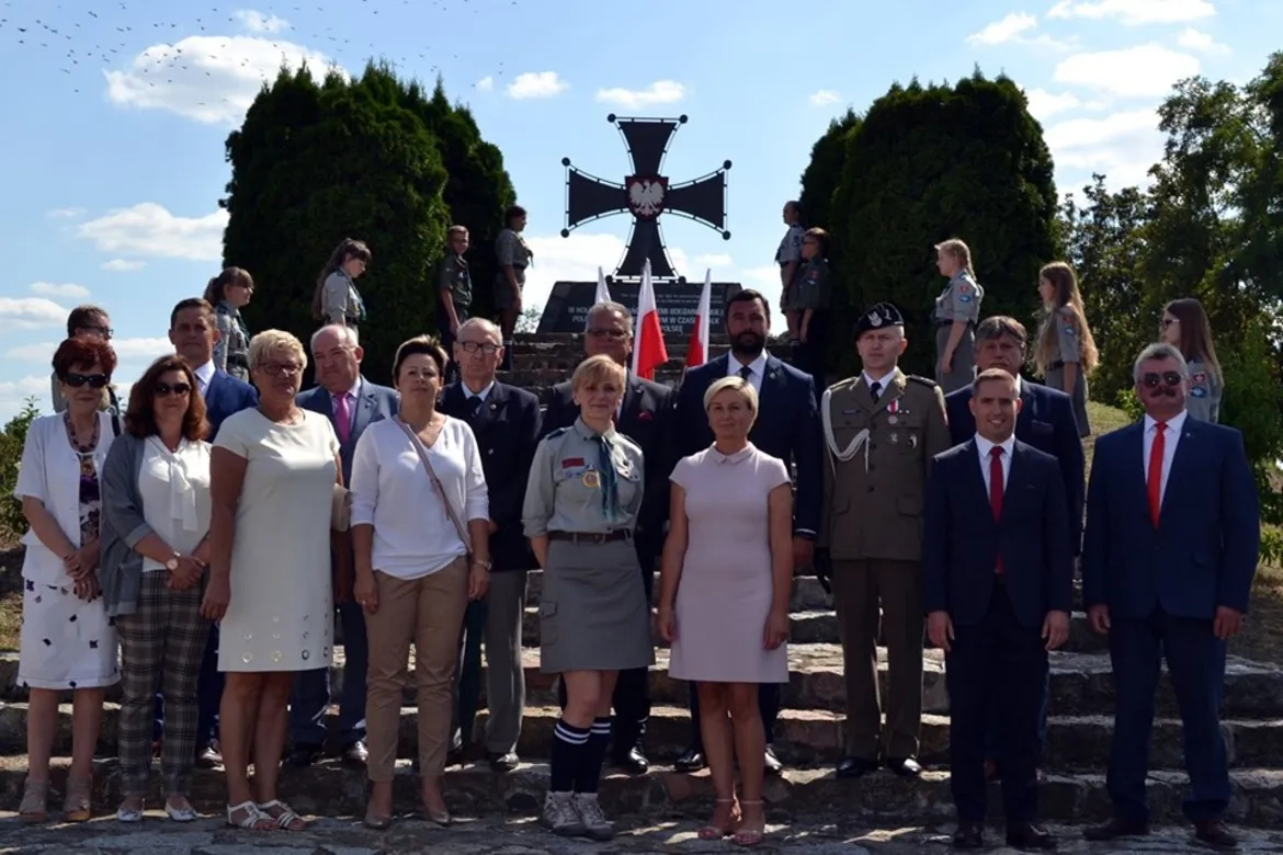 Grupa mieszkańców i osób w mundurach stoi na schodach przed pomnikiem w kształcie krzyża z orłem w centrum podczas Dni Bodzanowa 2019.