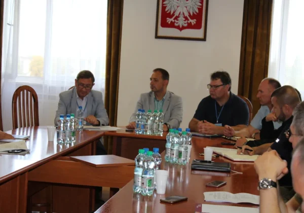 Kilka osób siedzi przy długim stole konferencyjnym podczas ćwiczenia obronnego Zielony Jar 2019, z butelkami wody i notatnikami na stole, a na ścianie wisi polskie godło – biały orzeł na czerwonej tarczy.