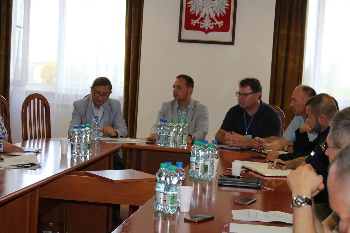 Kilka osób siedzi przy długim stole konferencyjnym podczas ćwiczenia obronnego Zielony Jar 2019, z butelkami wody i notatnikami na stole, a na ścianie wisi polskie godło – biały orzeł na czerwonej tarczy.