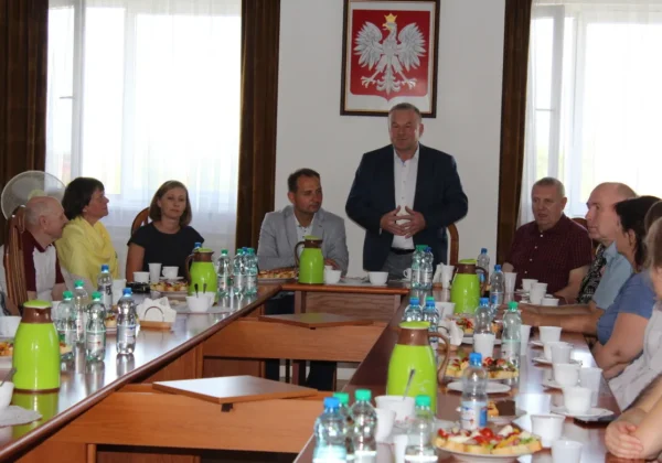 Potomkowie polskich zesłańców siedzą przy długim stole w sali konferencyjnej Powiatu Płockiego, słuchając przemówienia mężczyzny w garniturze; na ścianie wisi godło Polski.