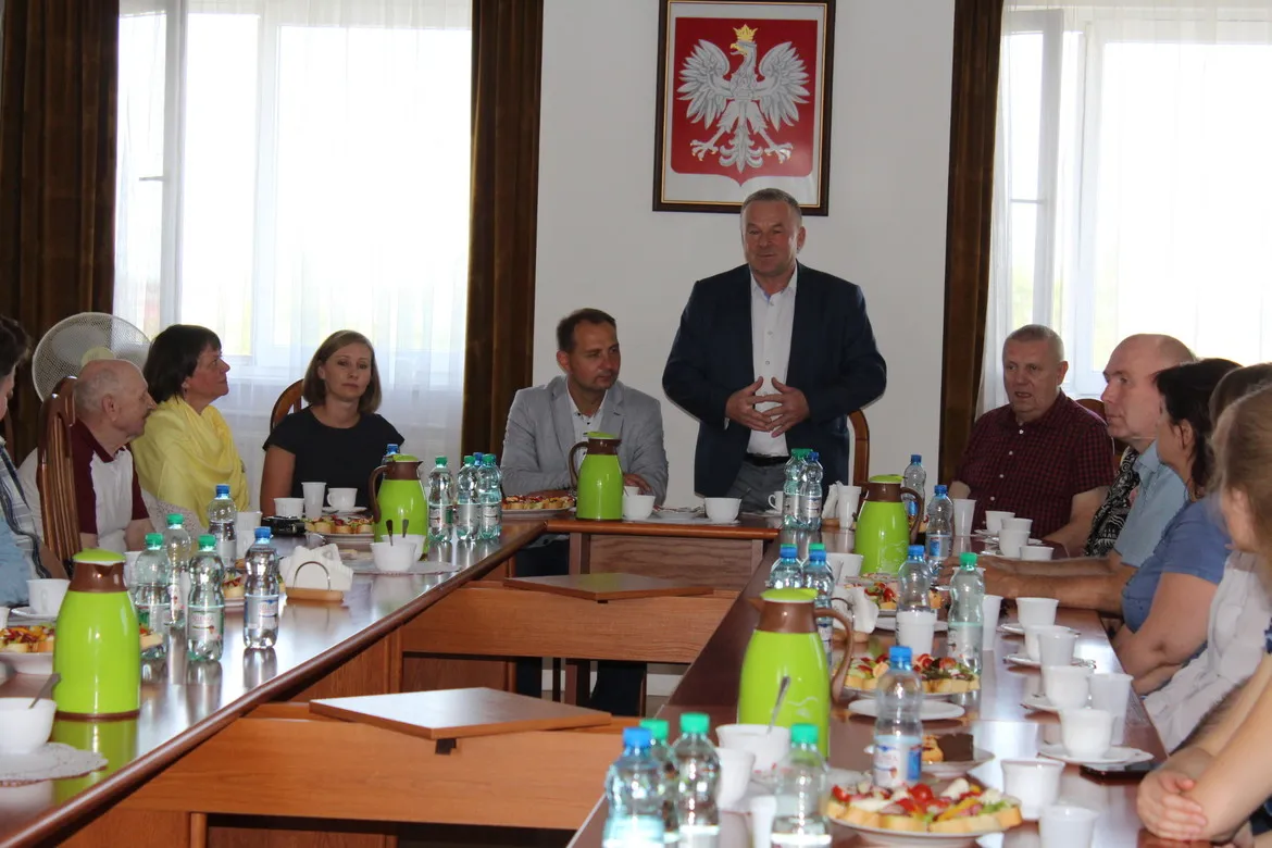 Potomkowie polskich zesłańców siedzą przy długim stole w sali konferencyjnej Powiatu Płockiego, słuchając przemówienia mężczyzny w garniturze; na ścianie wisi godło Polski.