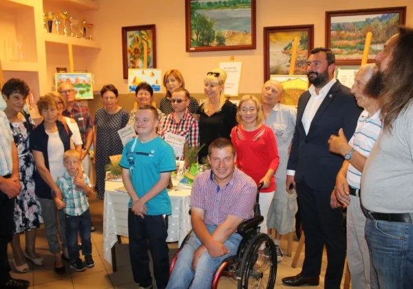 Grupa uczestników i gości podczas otwarcia nowych sal Środowiskowego Domu Samopomocy w Wyszogrodzie, zgromadzona wokół stołu z dyplomami i kwiatami, z mężczyzną na wózku inwalidzkim na pierwszym planie.