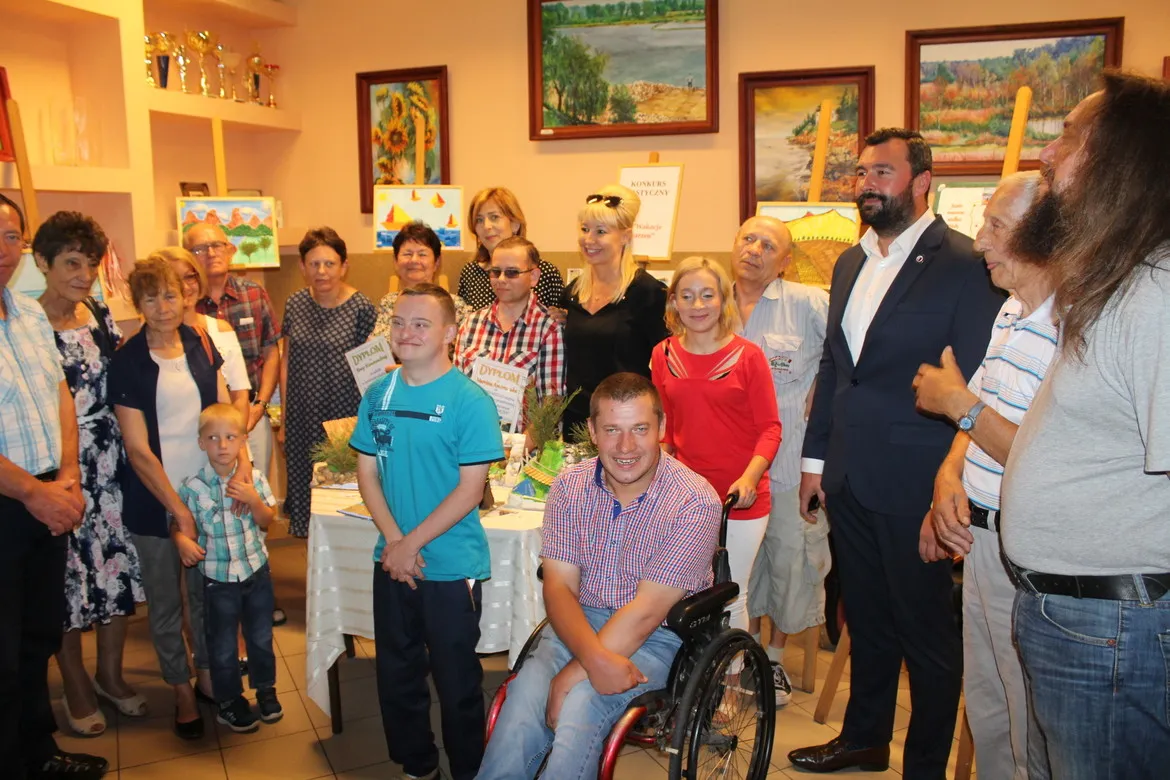 Grupa uczestników i gości podczas otwarcia nowych sal Środowiskowego Domu Samopomocy w Wyszogrodzie, zgromadzona wokół stołu z dyplomami i kwiatami, z mężczyzną na wózku inwalidzkim na pierwszym planie.