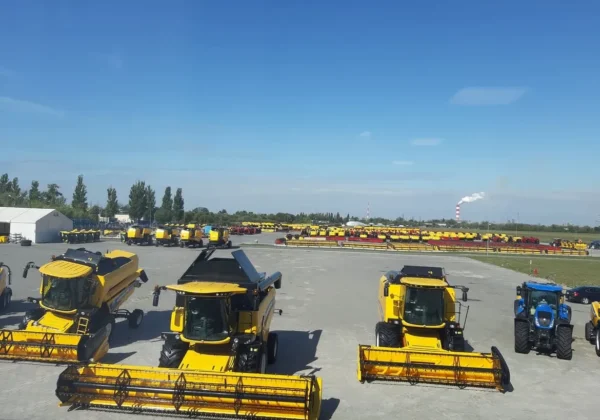 Na dużym placu pokazowym ustawiono liczne żółte maszyny rolnicze marki New Holland, w tym kombajny i traktory, a w tle widać nową halę i komin z dymem na błękitnym niebie.
