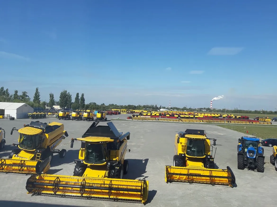 Na dużym placu pokazowym ustawiono liczne żółte maszyny rolnicze marki New Holland, w tym kombajny i traktory, a w tle widać nową halę i komin z dymem na błękitnym niebie.