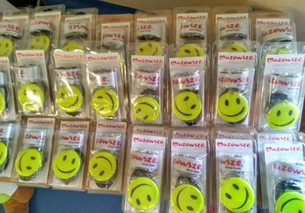 Na półce leży wiele żółtych, okrągłych emblematów ze śmiesznym uśmiechem, w opakowaniach z logo Mazowsze, będących częścią kampanii „Jesteś widoczny, jesteś bezpieczny – wyprawka dla pierwszoklasisty”.