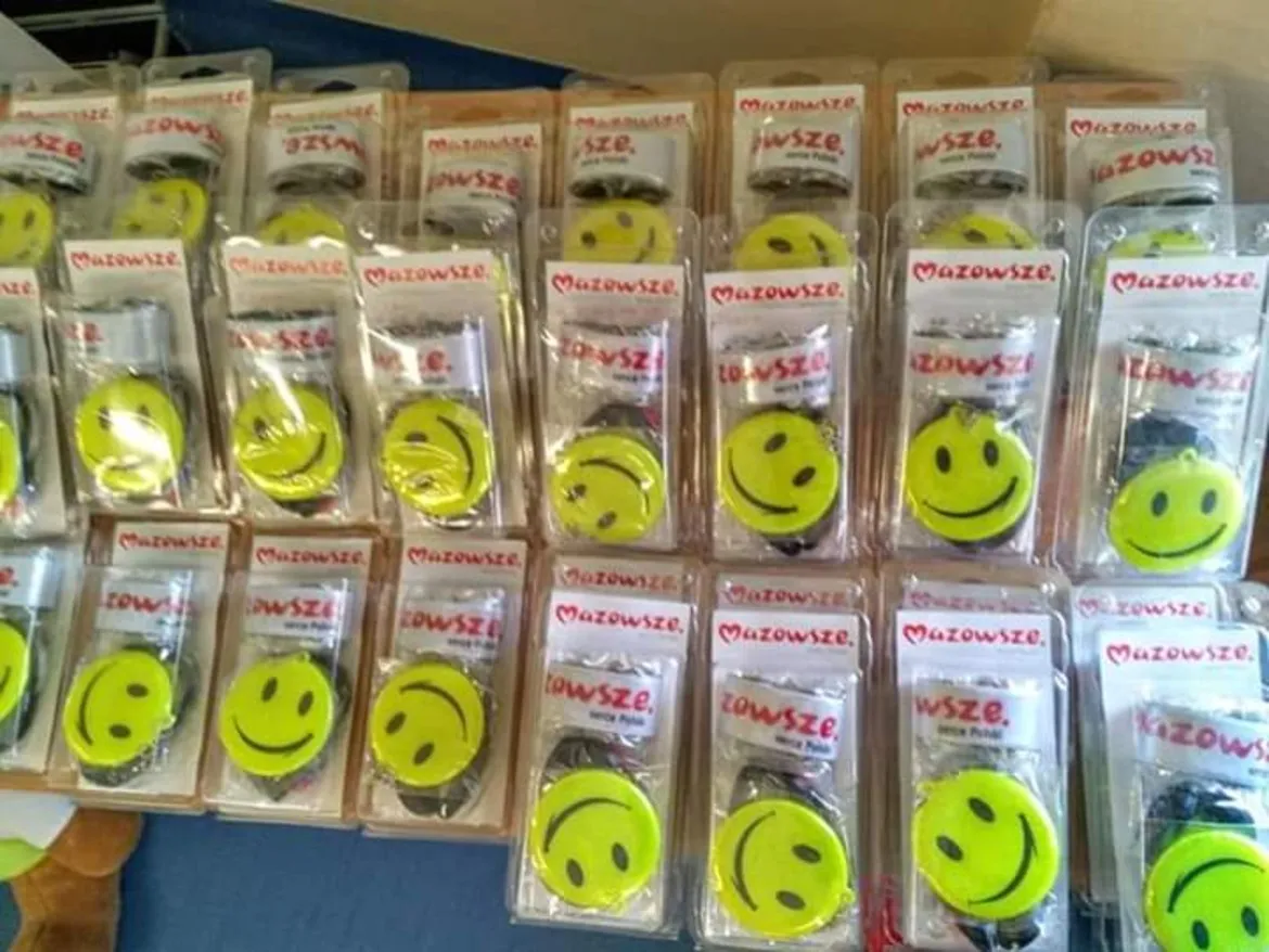 Na półce leży wiele żółtych, okrągłych emblematów ze śmiesznym uśmiechem, w opakowaniach z logo Mazowsze, będących częścią kampanii „Jesteś widoczny, jesteś bezpieczny – wyprawka dla pierwszoklasisty”.