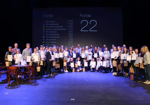 Grupa uczestników i organizatorów stoi na scenie z dyplomami i torbami upominków podczas zakończenia I Interaktywnego Turnieju Wiedzy o Powiecie Płockim i Ziemi Płockiej, na tle ekranu z wynikami rundy 22.