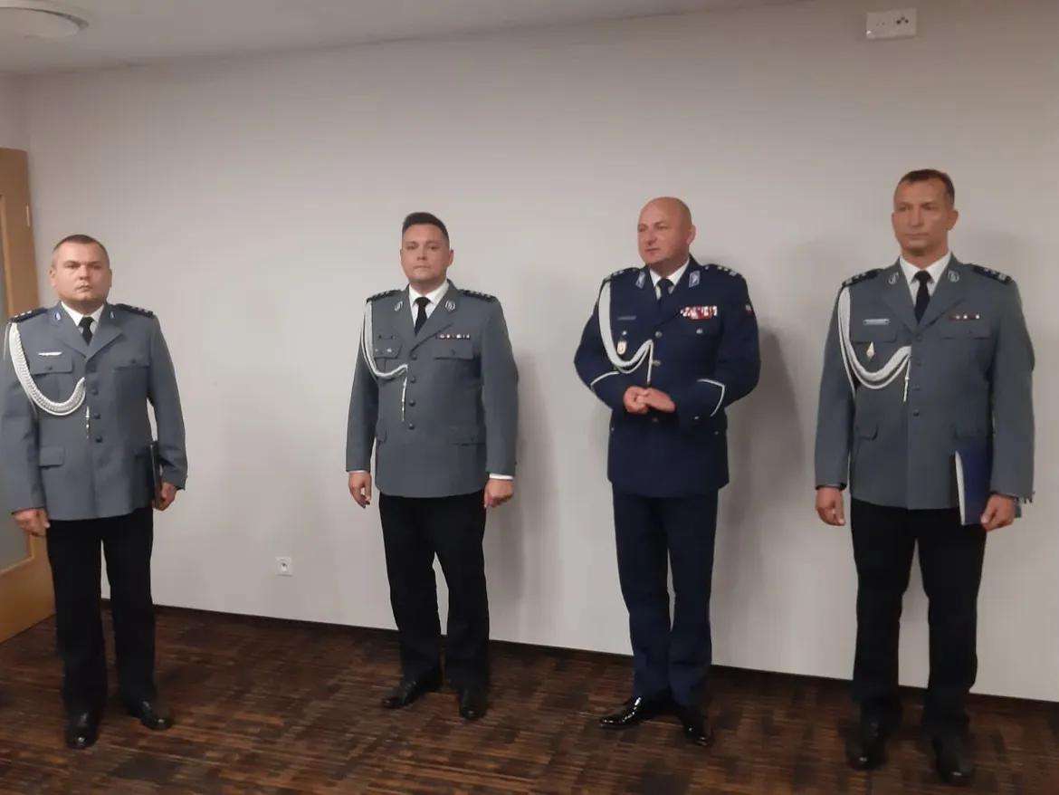 Czterech policjantów w mundurach stoi w pomieszczeniu z białą ścianą, podczas prezentacji nowego komendanta Komendy Miejskiej Policji w Płocku.