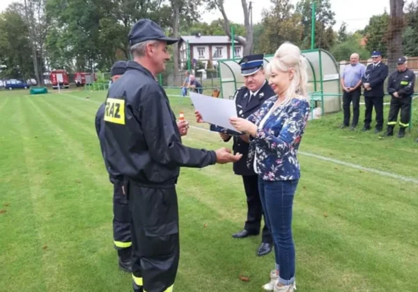 Strażak w czarnym kombinezonie z napisem STRAŻ odbiera dokument od kobiety w kwiecistej marynarce podczas ceremonii wręczania czeków dla strażaków na boisku.