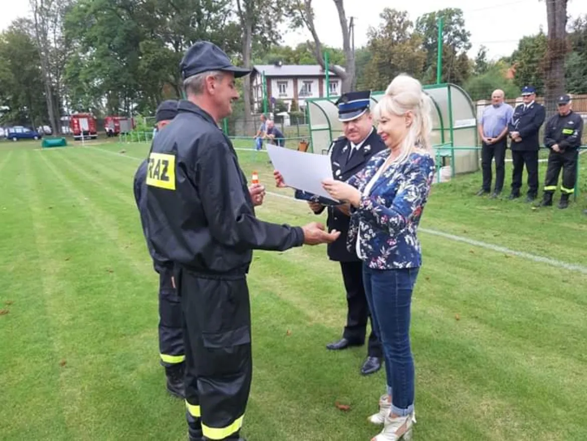 Strażak w czarnym kombinezonie z napisem STRAŻ odbiera dokument od kobiety w kwiecistej marynarce podczas ceremonii wręczania czeków dla strażaków na boisku.