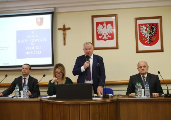 Podczas X Sesji Rady Powiatu w Płocku mężczyzna w garniturze przemawia do mikrofonu stojąc przy stole, obok siedzą dwie osoby, a w tle widać projekcję ekranu z napisem i herby na ścianie.