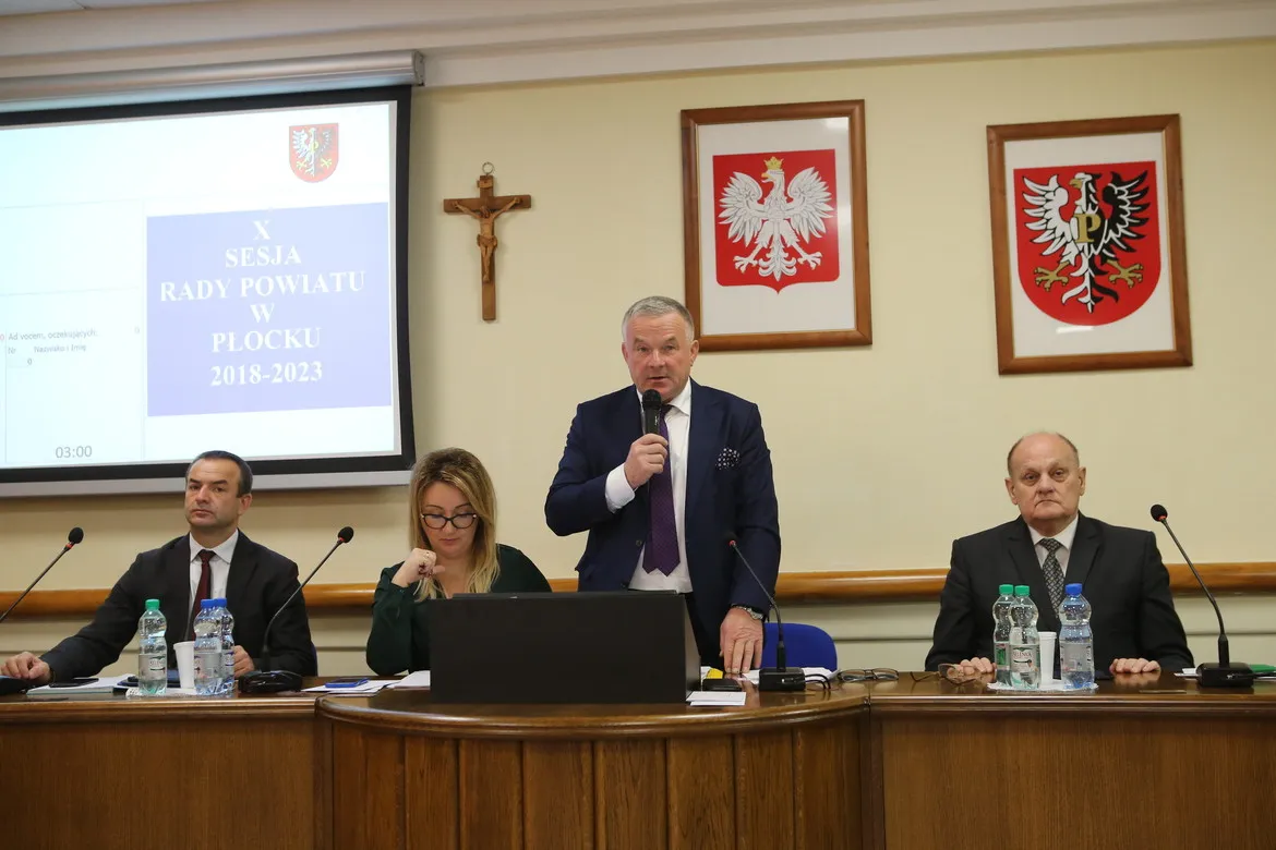 Podczas X Sesji Rady Powiatu w Płocku mężczyzna w garniturze przemawia do mikrofonu stojąc przy stole, obok siedzą dwie osoby, a w tle widać projekcję ekranu z napisem i herby na ścianie.
