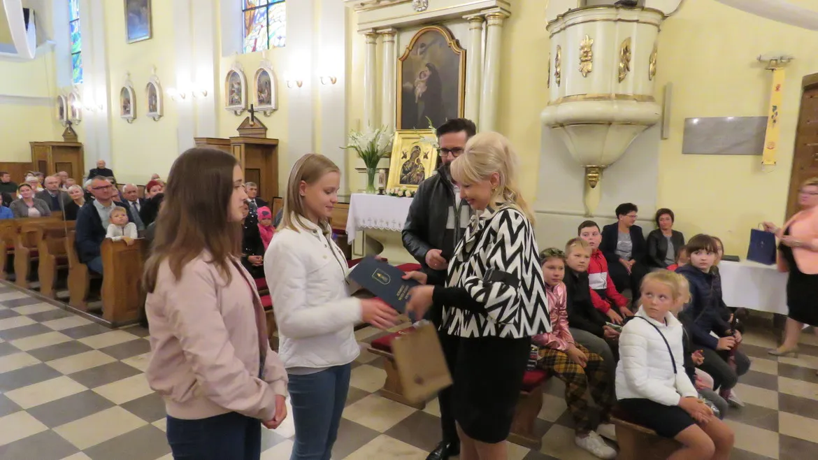 Podczas Święta Stanisława Kostki w Drobinie w kościele kobieta wręcza dyplom dwóm dziewczynkom stojącym na środku, podczas gdy zebrani mieszkańcy obserwują.