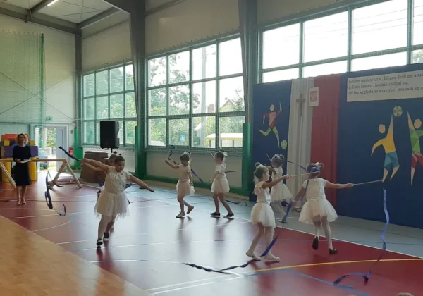 Grupa dziewczynek w białych tutu tańczy z wstążkami w sali gimnastycznej podczas szkolnego pokazu.