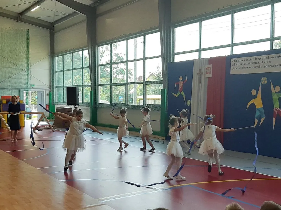 Grupa dziewczynek w białych tutu tańczy z wstążkami w sali gimnastycznej podczas szkolnego pokazu.