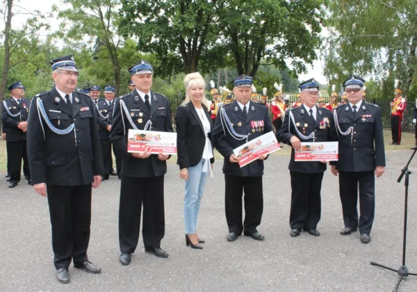 Uroczystość 70-lecia OSP Miszewko Strzałkowskie: grupa strażaków w mundurach z odznaczeniami i kobieta w czarnym żakiecie trzymają certyfikaty, stojąc na plenerowym placu z orkiestrą w tle.