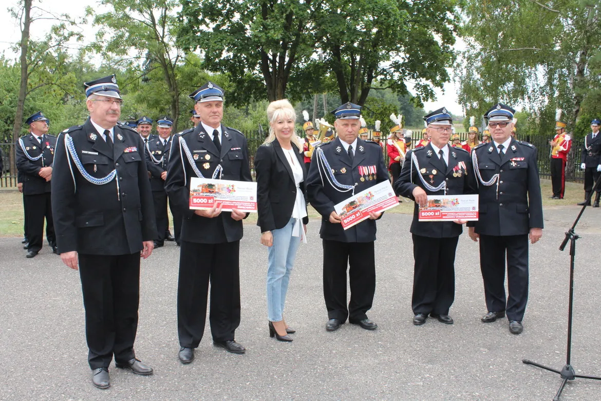 Uroczystość 70-lecia OSP Miszewko Strzałkowskie: grupa strażaków w mundurach z odznaczeniami i kobieta w czarnym żakiecie trzymają certyfikaty, stojąc na plenerowym placu z orkiestrą w tle.