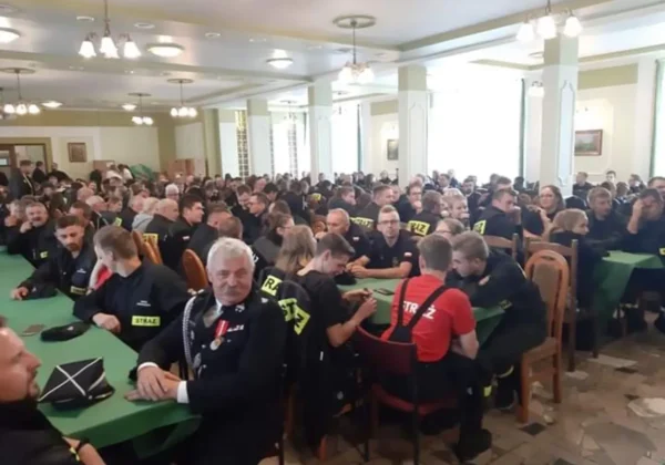 Duża grupa druhów Ochotniczej Straży Pożarnej siedzi przy zielonych stołach w dużej sali podczas Powiatowego Konkursu Ekologicznego.