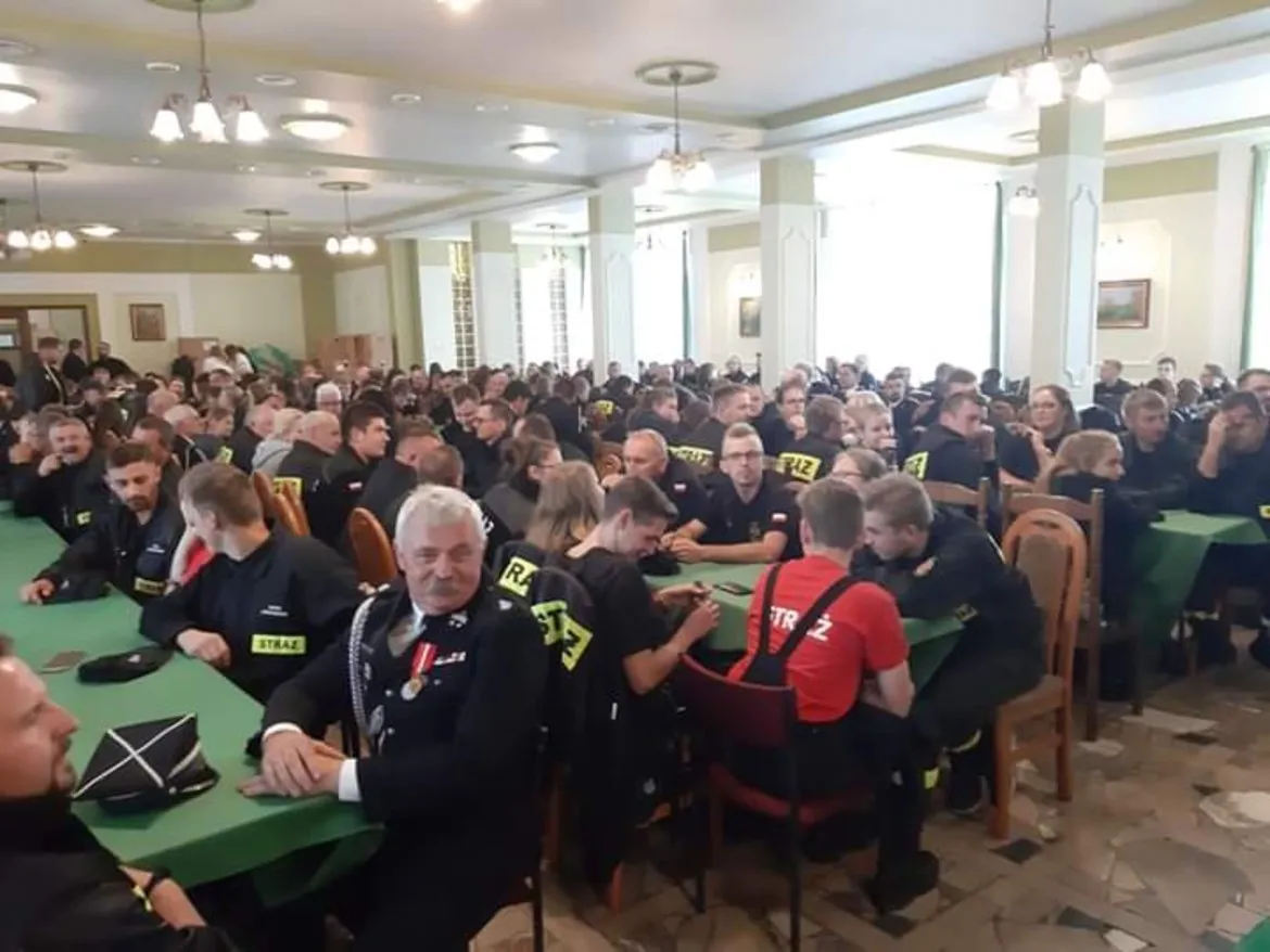 Duża grupa druhów Ochotniczej Straży Pożarnej siedzi przy zielonych stołach w dużej sali podczas Powiatowego Konkursu Ekologicznego.