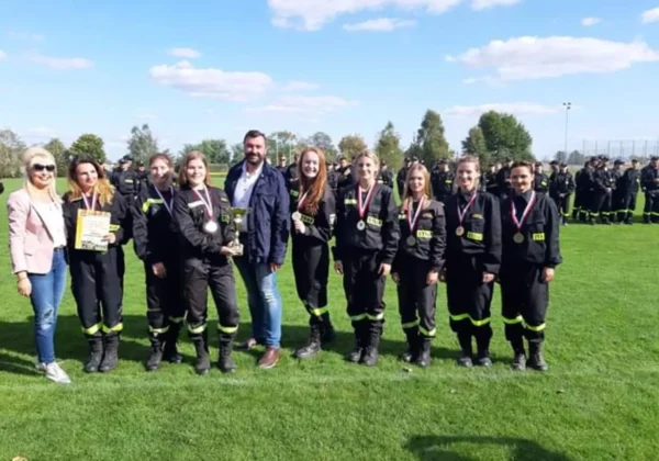 Grupa strażaków w czarnych mundurach z odblaskami i medalami stoi na zielonej trawie podczas XI Powiatowych Zawodów Sportowo-Pożarniczych, niektórzy trzymają puchar i certyfikat.