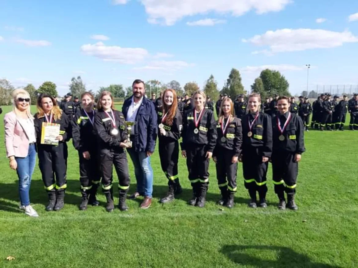 Grupa strażaków w czarnych mundurach z odblaskami i medalami stoi na zielonej trawie podczas XI Powiatowych Zawodów Sportowo-Pożarniczych, niektórzy trzymają puchar i certyfikat.