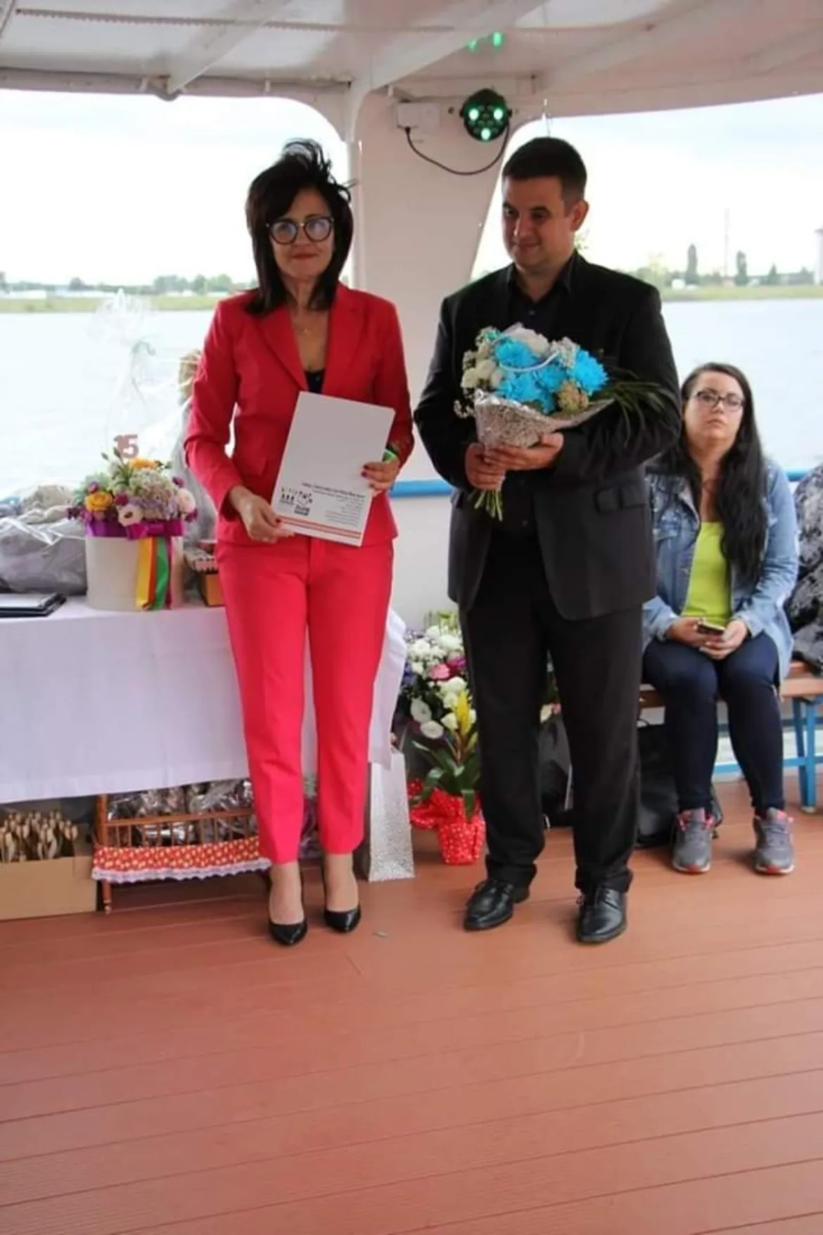 Na tarasie nad jeziorem kobieta w czerwonym garniturze wręcza dokument mężczyźnie, który trzyma bukiet kwiatów, podczas jubileuszu 15-lecia Funduszu Lokalnego Ziemi Płockiej – Młodzi Razem i Działaj Lokalnie.