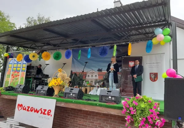 Scena plenerowa podczas Dnia Pieczonego Ziemniaka w DPS Goślice: kobieta z mikrofonem i mężczyzna z teczką stoją na scenie, otoczeni kolorowymi ozdobami, balonami i kwiatami, przy banerze Mazowsze – serce Polski.