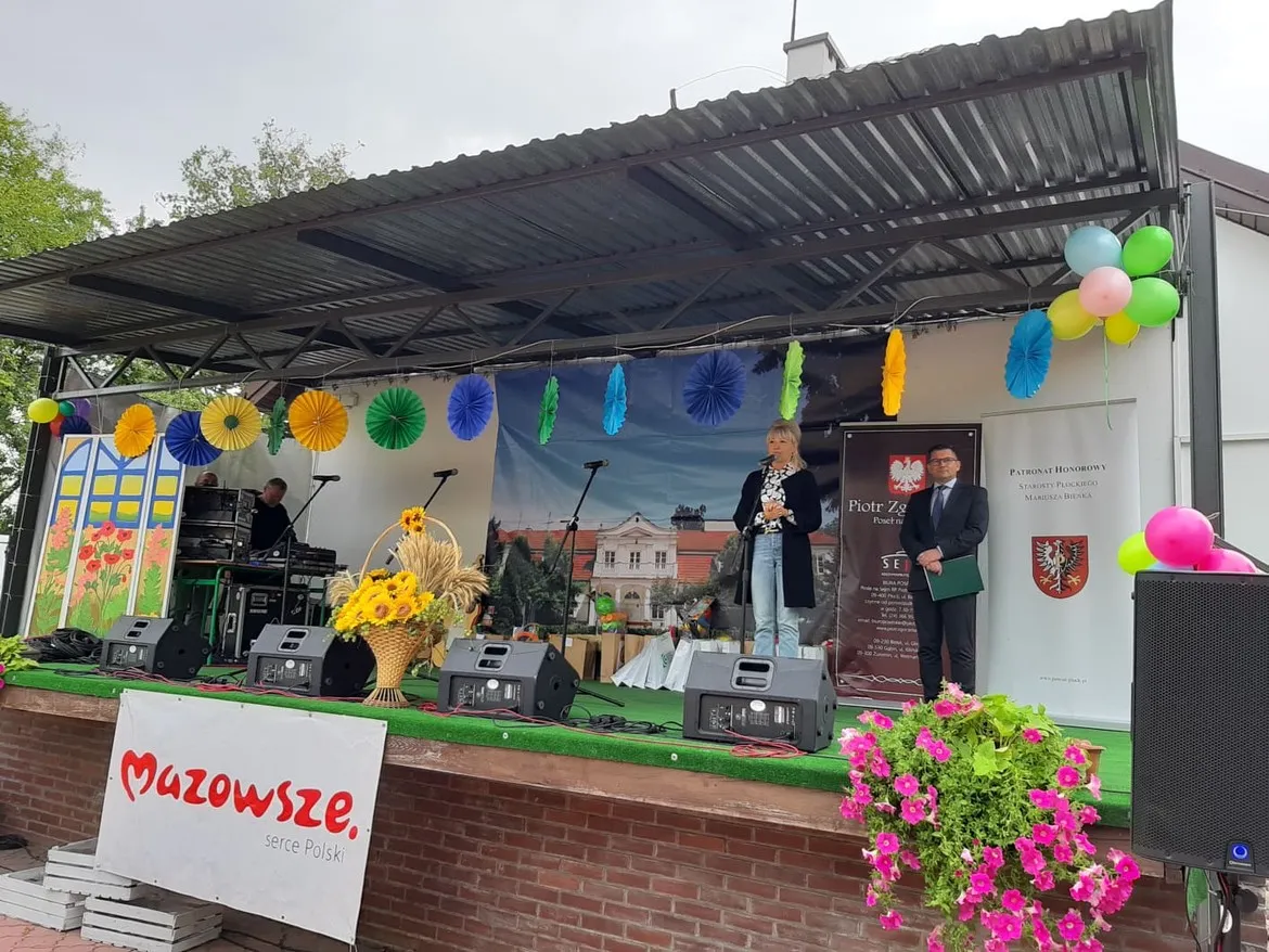 Scena plenerowa podczas Dnia Pieczonego Ziemniaka w DPS Goślice: kobieta z mikrofonem i mężczyzna z teczką stoją na scenie, otoczeni kolorowymi ozdobami, balonami i kwiatami, przy banerze Mazowsze – serce Polski.