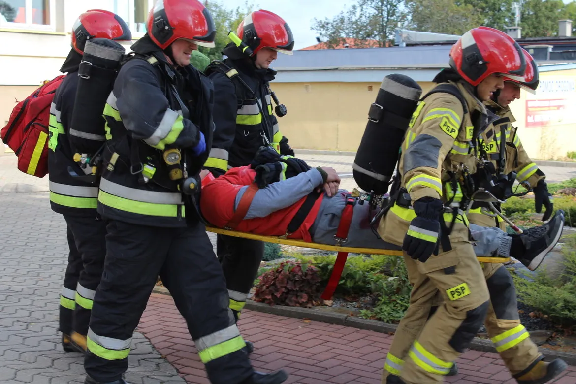 Grupa strażaków w hełmach i ubraniach ochronnych niesie na noszach rannego podczas ćwiczeń obronnych Zielony Jar 2019.