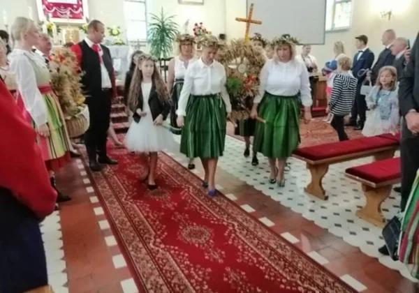Uczestnicy Dożynkowego Święta w Powiecie Płockim w kościele, ubrani w tradycyjne ludowe stroje z zielonymi spódnicami, idą alejką.