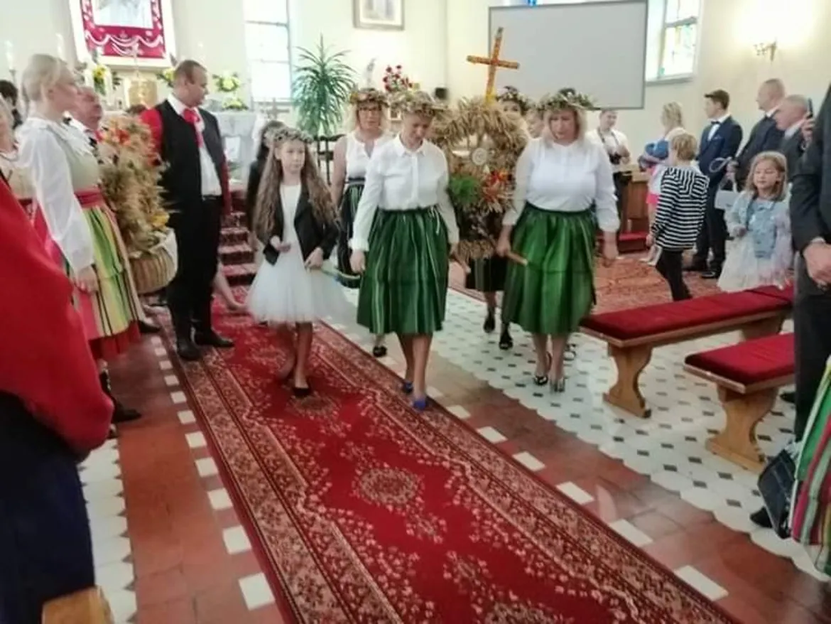 Uczestnicy Dożynkowego Święta w Powiecie Płockim w kościele, ubrani w tradycyjne ludowe stroje z zielonymi spódnicami, idą alejką.