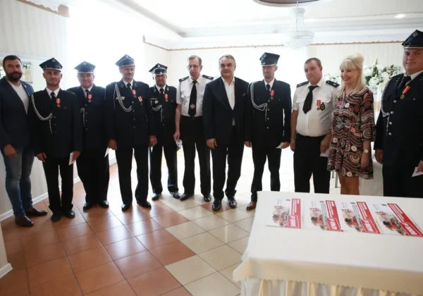 Grupa osób, w tym strażacy w mundurach z odznaczeniami i kilka osób w cywilnych ubraniach, stoi razem w sali podczas wizyty Prezesa Zarządu Głównego ZOSP RP w powiecie płockim, obok stołu z ulotkami.