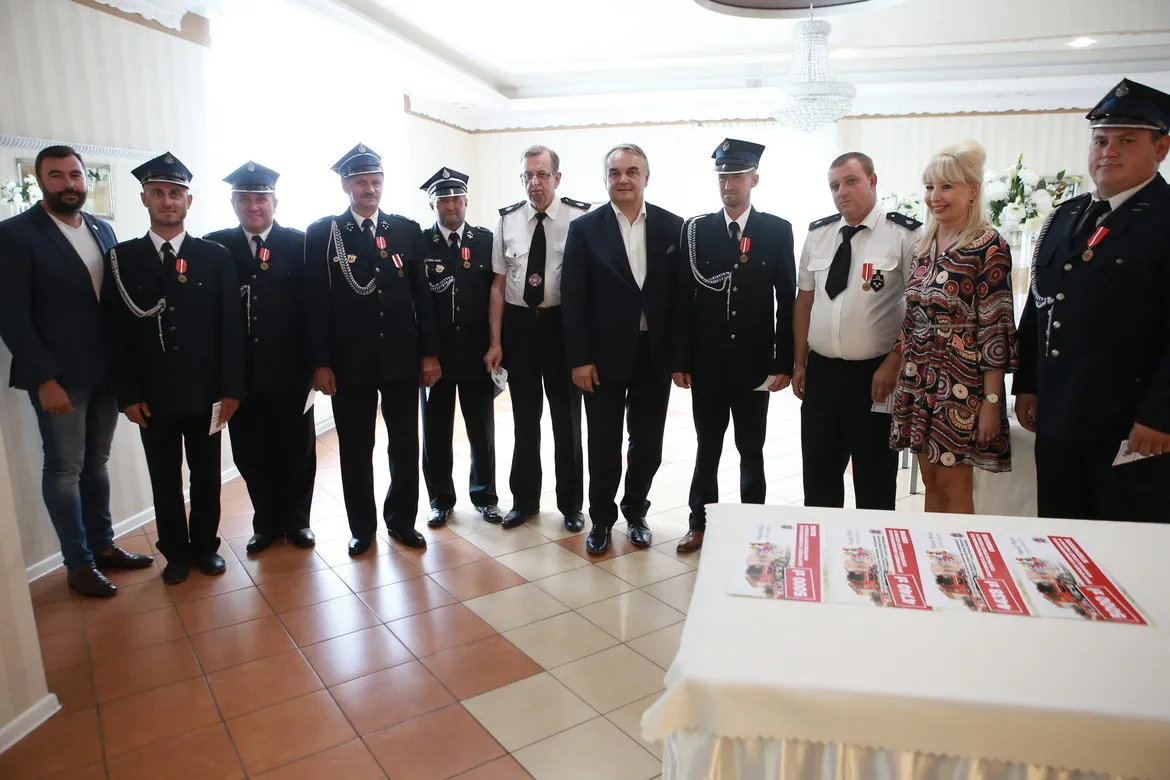 Grupa osób, w tym strażacy w mundurach z odznaczeniami i kilka osób w cywilnych ubraniach, stoi razem w sali podczas wizyty Prezesa Zarządu Głównego ZOSP RP w powiecie płockim, obok stołu z ulotkami.