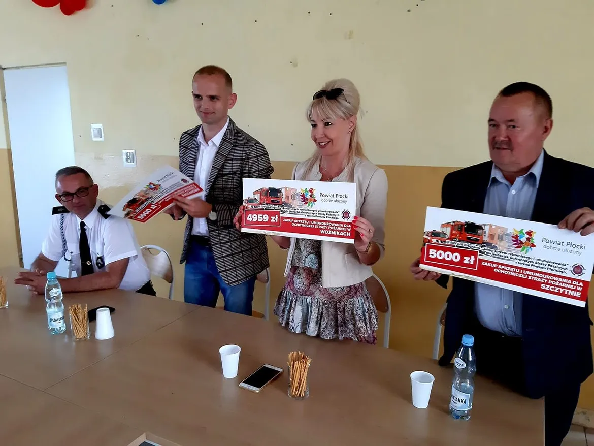 Czterech przedstawicieli Powiatu Płockiego siedzi przy stole i trzyma plakietki z kwotami 4959 zł i 5000 zł, symbolizujące kolejne wsparcie dla OSP w Radzanowie, Woźnikach i Szczytnie.