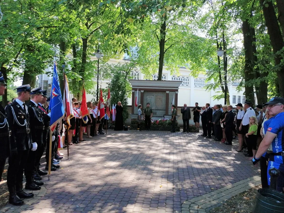 Uczestnicy obchodów 80. rocznicy wybuchu II wojny światowej w Gąbinie stoją po obu stronach alejki przed centralnym pomnikiem, trzymając flagi.